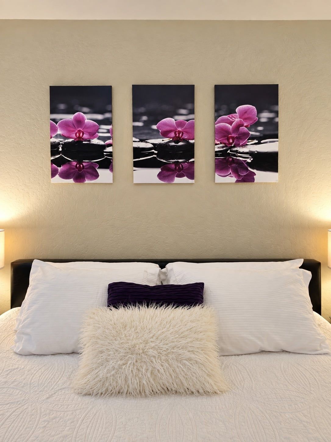 A bedroom with a bed, white and purple pillows, and three wall-mounted photographs of pink orchids.