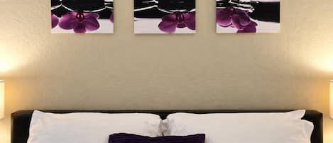 Un dormitorio con cama, almohadas blancas y púrpuras, y tres fotografías colgadas en la pared de orquídeas rosadas.