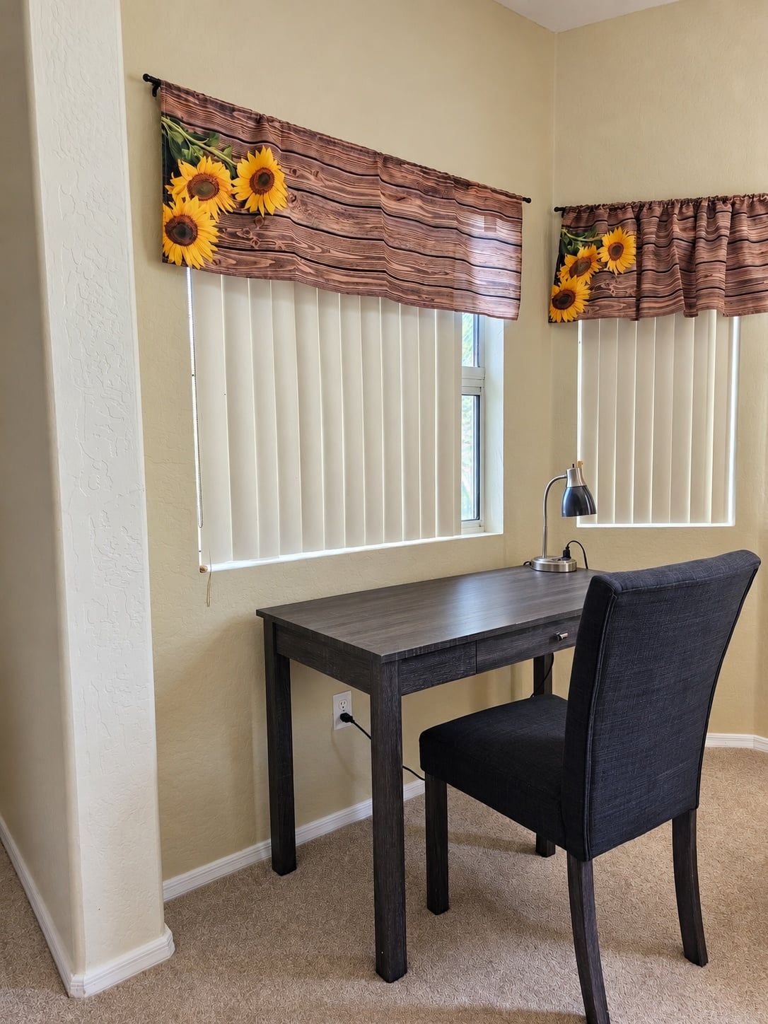 A room with a dark wooden desk, a black chair, and two windows with brown curtains featuring sunflower designs.