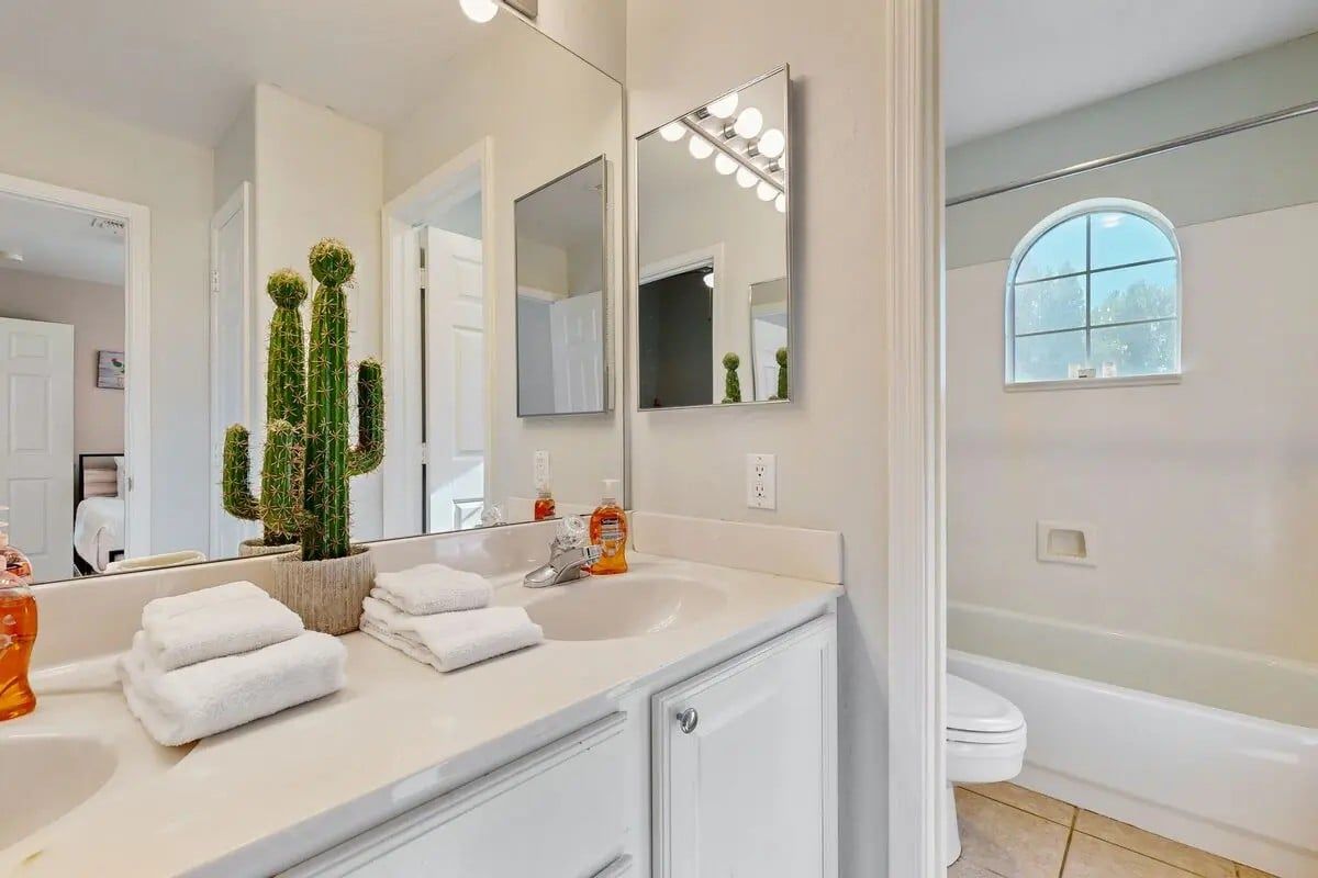 A bathroom with a white vanity, a large mirror with lights, a cactus decoration, and a window.