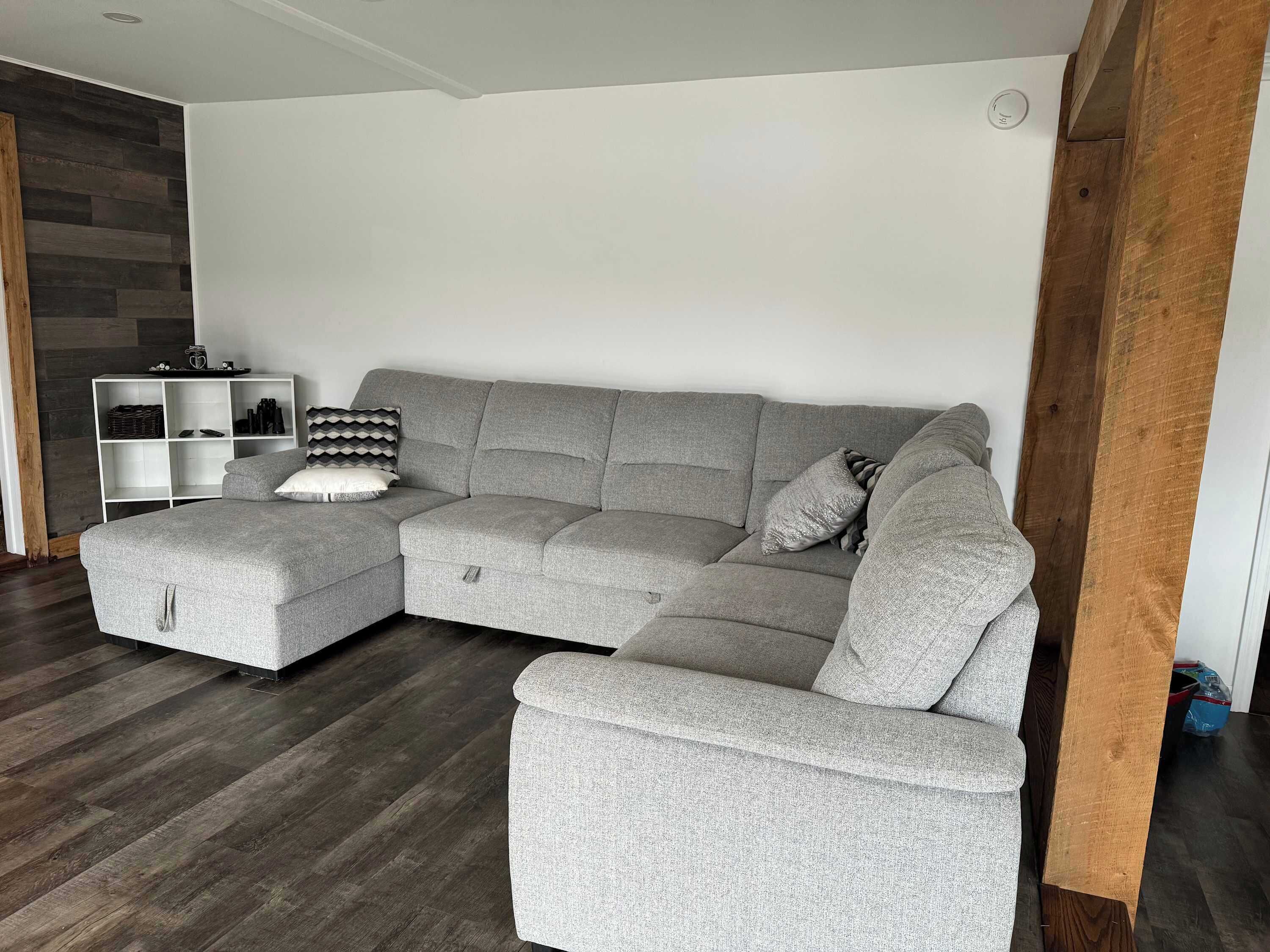 Ein graues Ecksofa mit Stauraum-Hocker, eine weiße Regaleinheit und eine Holz-Akzentwand.