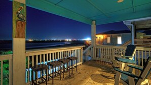 A wooden deck with chairs and tables, overlooking a beach at night. - La Sirena by Avantstay Communal Pool + Covered Lounge + Marsh Views + Near Port A Beach (Port Aransas)
