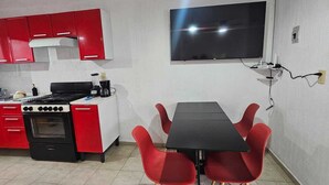 Une cuisine compacte avec des meubles rouges, une cuisinière noire et une table à manger noire avec des chaises rouges. Un téléviseur à écran plat est fixé au mur au-dessus de l’espace repas.