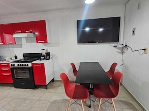 A compact kitchen with red cabinets, a black stove, and a black dining table with red chairs. A flat-screen TV is mounted on the wall above the dining area.