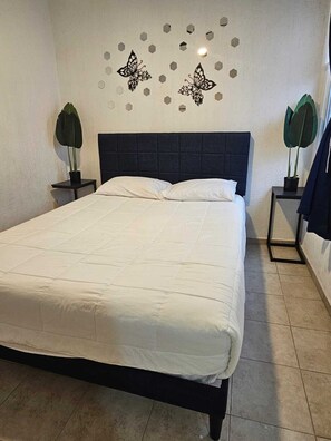 Un lit bien fait dans une chambre avec une tête de lit sombre, deux tables de chevet avec des lampes et des œuvres d’art décoratives accrochées au mur.