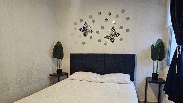 Un lit bien fait, avec une literie blanche, dans une pièce dont la tête de lit est de couleur sombre. On y trouve deux tables de chevet ornées de plantes en pot, ainsi qu’une décoration murale représentant des papillons et des hexagones.