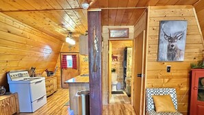 Een gezellig blokhutinterieur met houten wanden en vloeren, een kitchenette, een badkamer en een kleine zithoek met een gestreept kussen.