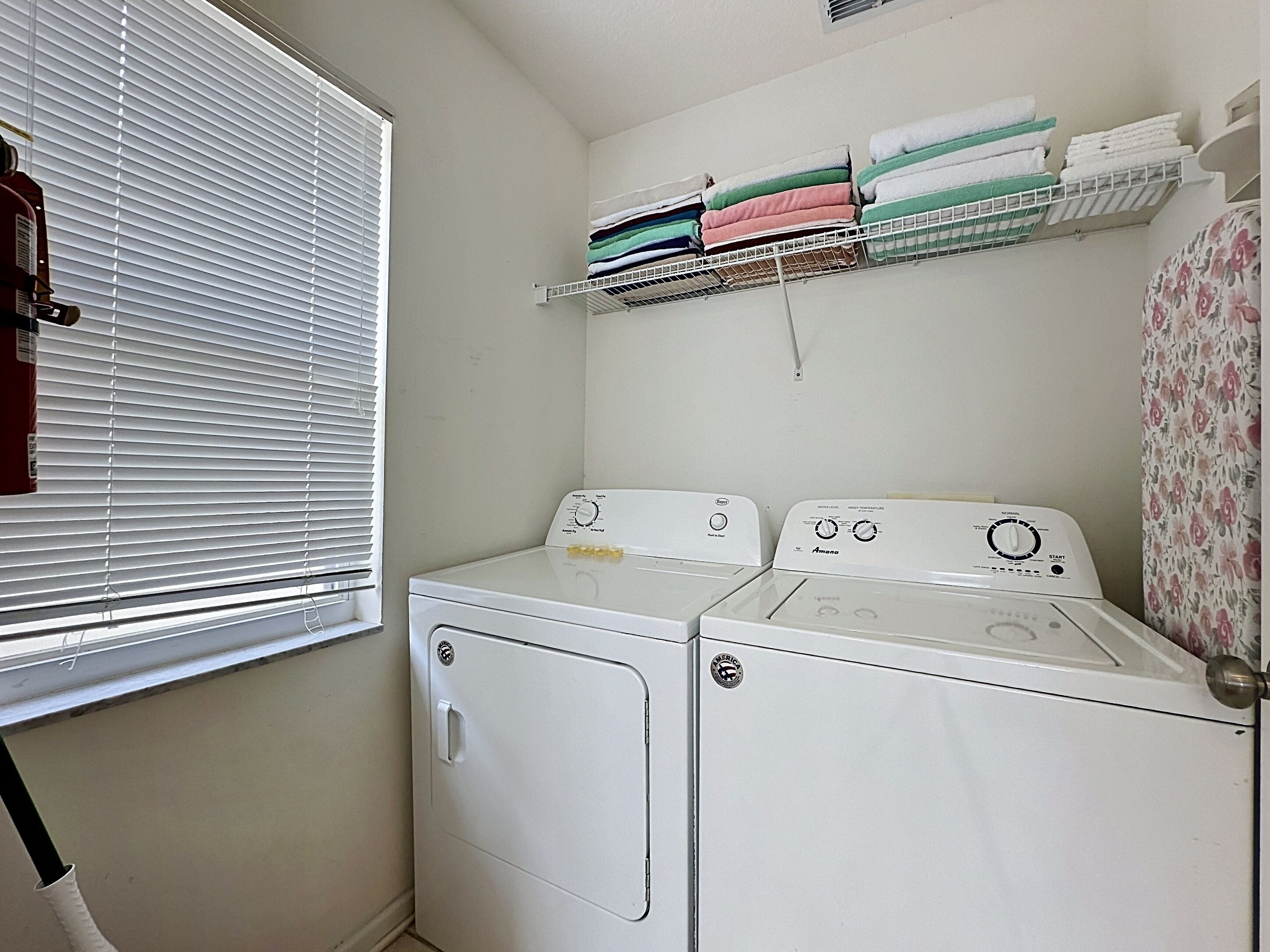 A laundry room with two white washing machines, a window with blinds, and a shelf with folded towels.