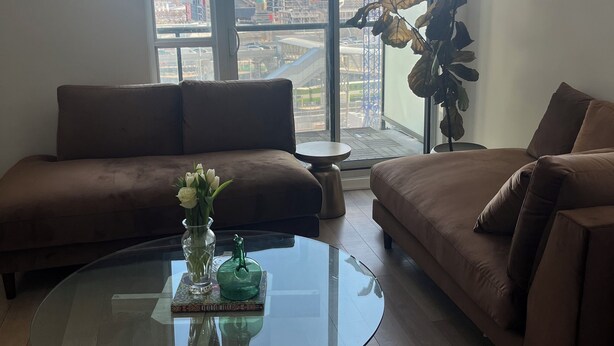 Ein modernes Wohnzimmer mit einem Glastisch, zwei braunen Sofas und Blick auf eine Baustelle durch große Fenster.