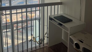 Ein Zimmer mit großem Fenster und Blick auf eine Baustelle, einem weißen Schreibtisch mit einem Laptop, einer kleinen Pflanze und einem gerahmten Bild an der Wand.