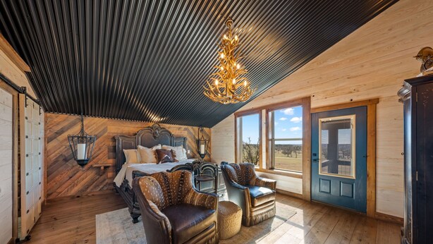 Ein Schlafzimmer mit einem Bett, zwei Sesseln, einer Kristallleuchter, einer Holzbalkendecke und Blick ins Freie.