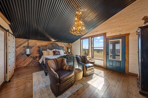 Een slaapkamer met een bed, twee fauteuils, een kroonluchter, een houten plafond en uitzicht op het landschap.