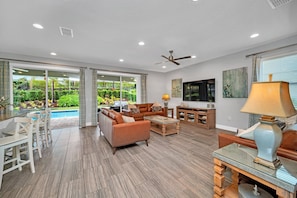 A spacious living room with a pool view, a ceiling fan, and a TV mounted on the wall. - Modern 6BR Villa / Games Room/ Private Pool & Spa/10 Min to Disney (Kissimmee)