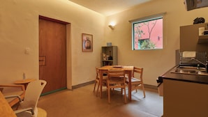 Een kleine keuken met een eethoek, een houten deur, een lijst met een afbeelding en een raam met uitzicht op groen.