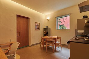 A compact kitchen with a dining area, a wooden door, a framed picture, and a window with a view of greenery. - Casona Soledad - Margarita (Oaxaca de Juárez)