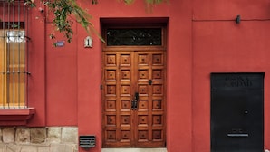 Een rood gebouw met een grote houten dubbele deur, een zwarte deur rechts en een raam met een metalen tralies links.