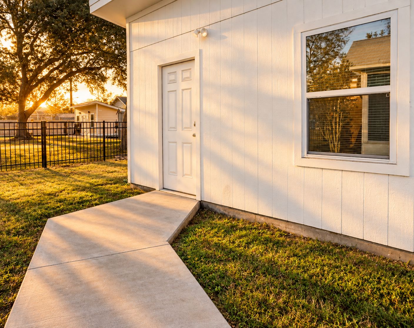 A residential house with a front door, a window, a concrete pathway, and a fenced yard.