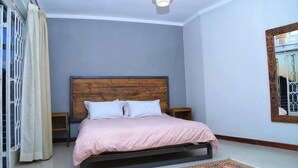 A bedroom with a wooden headboard, a single bed with a pink bedspread, two bedside tables, a patterned rug, and a large mirror with a decorative frame. - Kololo 3BR with Garden, 500m to Acacia Mall (Kampala)