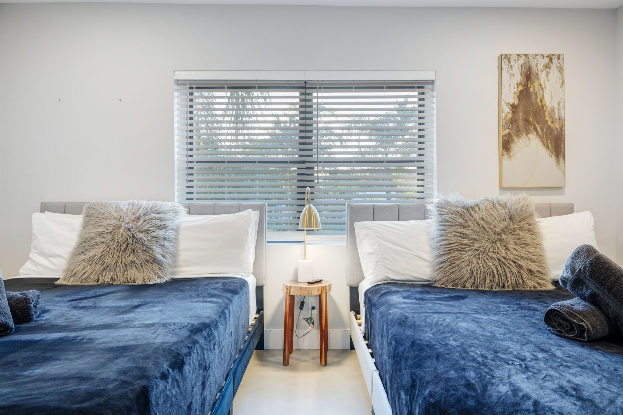 A room with two beds, a small wooden table, a lamp, and a window with blinds.