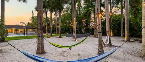 A hammock on a sandy beach with palm trees and a building in the background.