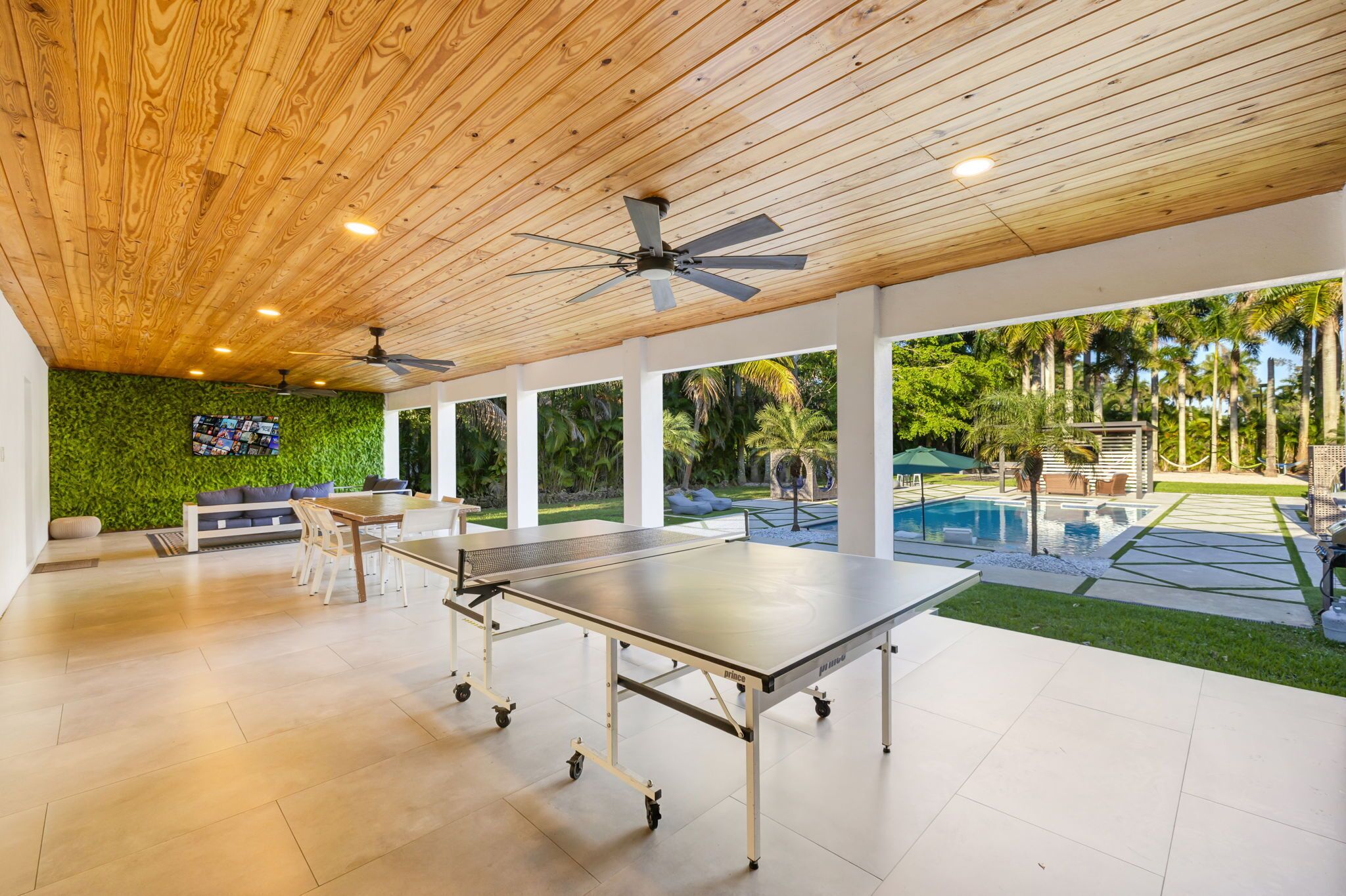 An indoor space with a ping pong table, ceiling fans, a TV, and a pool area visible through large windows.