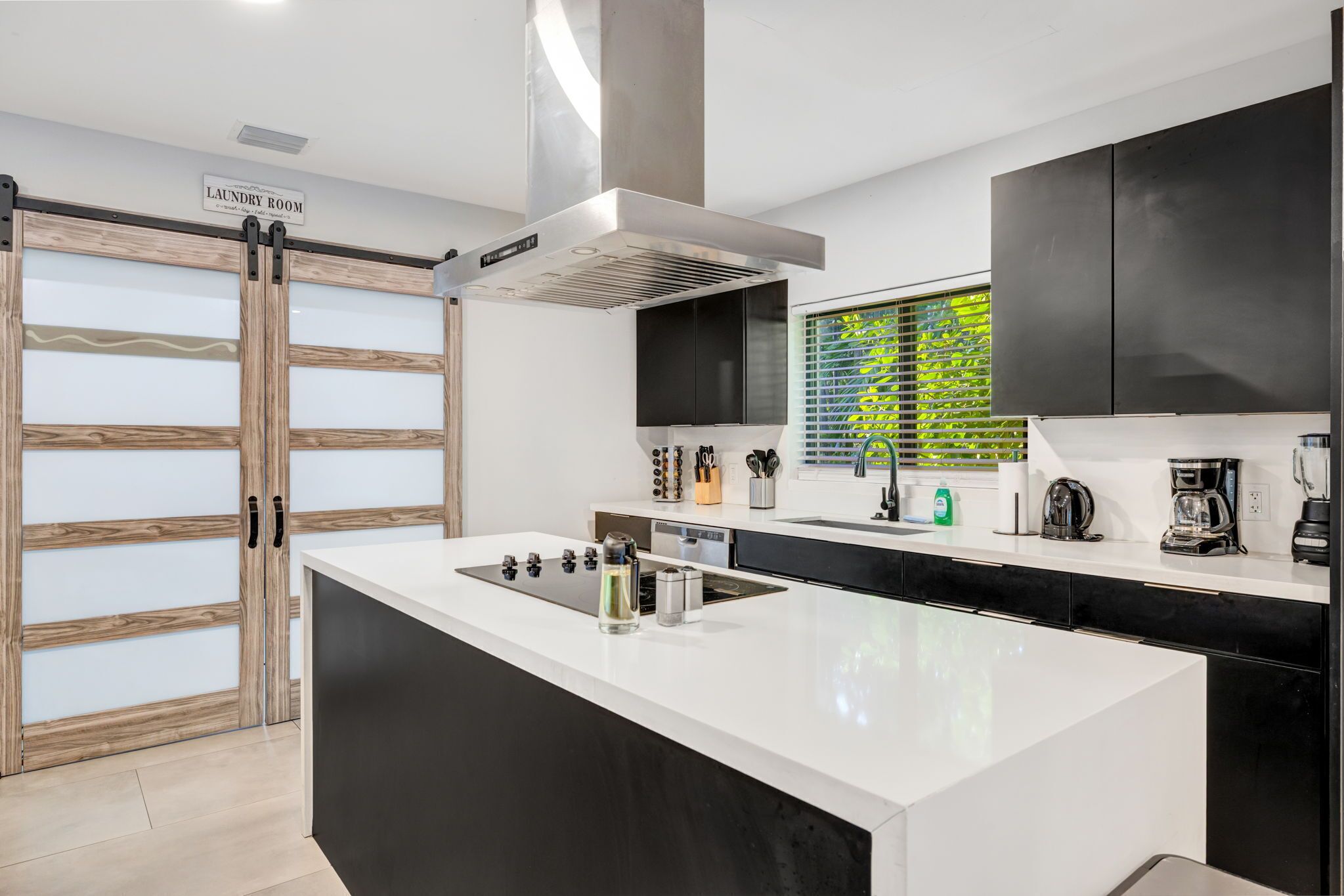Een moderne keuken met een centraal eiland, zwarte kastjes, een roestvrijstalen afzuigkap en een bordje dat de weg wijst naar de wasserette.