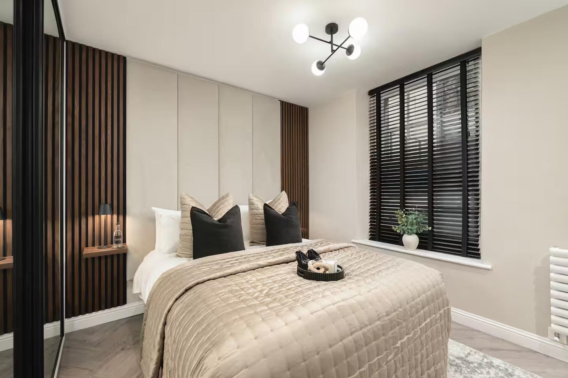 A bedroom with a bed, a bedside table, a window with blinds, and a ceiling light fixture.