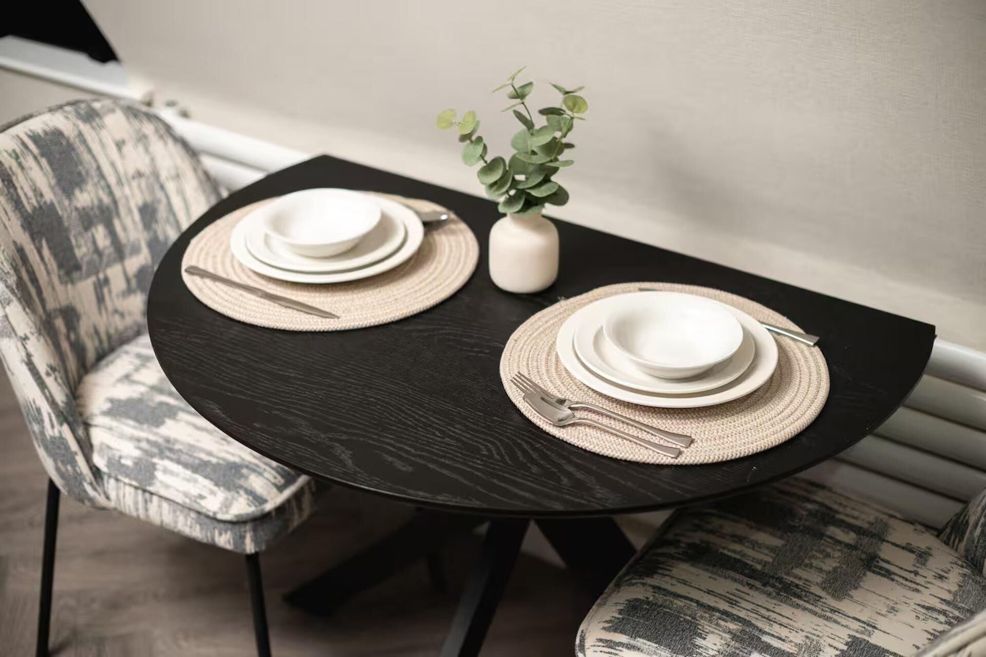 A dining table with two sets of white plates and cutlery, accompanied by woven placemats and a small potted plant.
