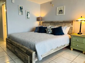 A bedroom with a wooden bed frame, a grey bedspread, and multiple pillows. There is a green nightstand with a lamp, and artwork on the walls. - DAYTONA BEACHFRONT Condo, Amazing OCEAN Views, Heated Pool, Hot Tub, King Bed (Daytona Beach)