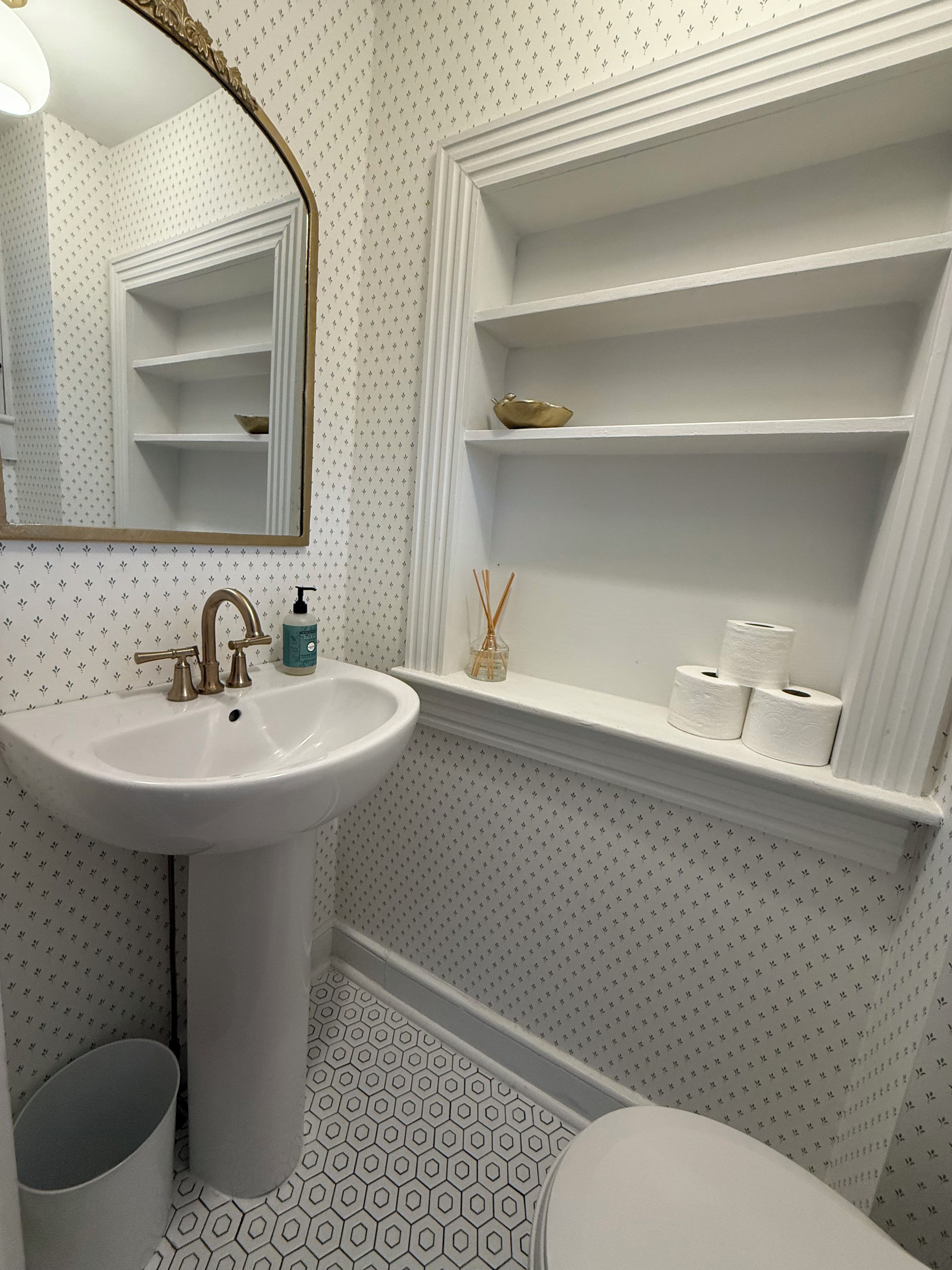 Et badeværelse med en hvid vask, et spejl, et toilet og en hylde med toiletartikler.