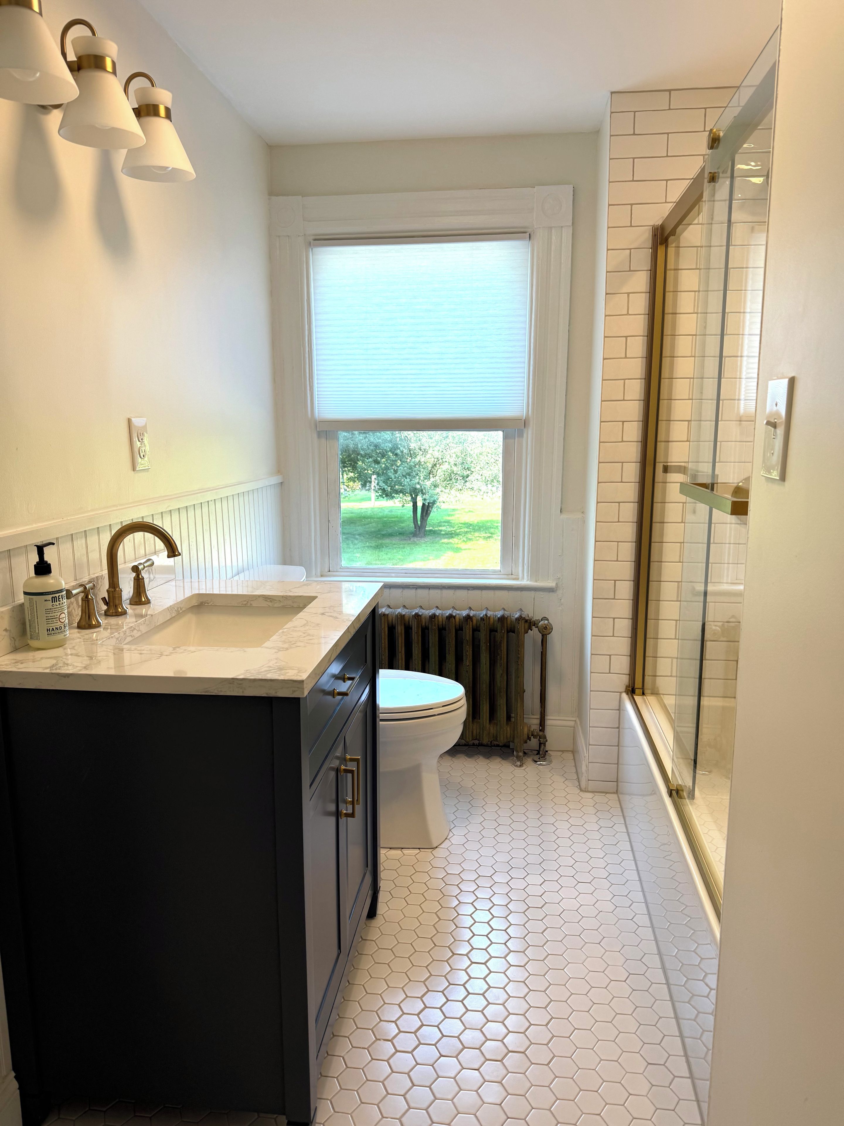 A bathroom with a white toilet, a sink with a marble countertop, a shower with a glass door, and a window with a view of greenery.