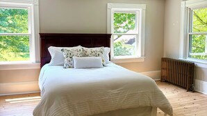 A bedroom with a large bed, two windows, a radiator, and a ceiling fan.