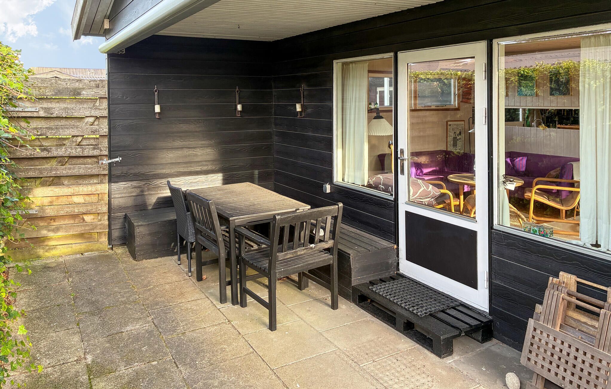A wooden cabin with a patio area featuring a table and chairs, a window with curtains, and a door.