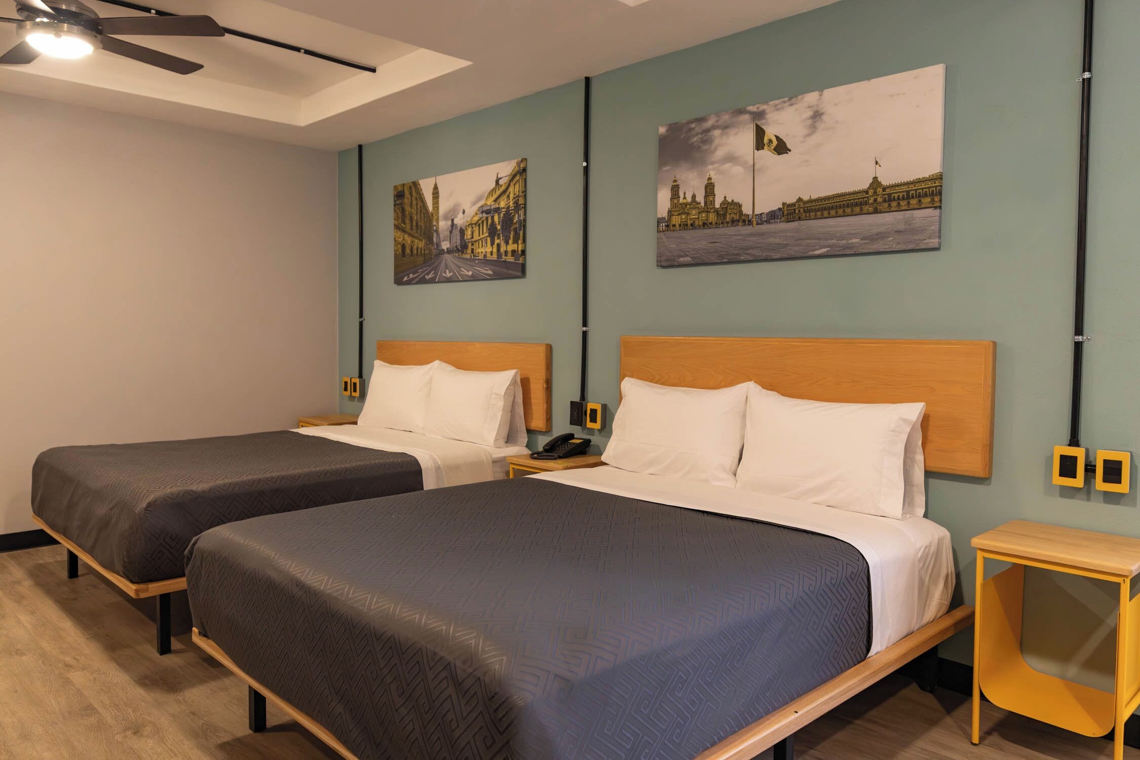 A hotel room with two beds, a ceiling fan, artwork on the walls, and a wooden nightstand.