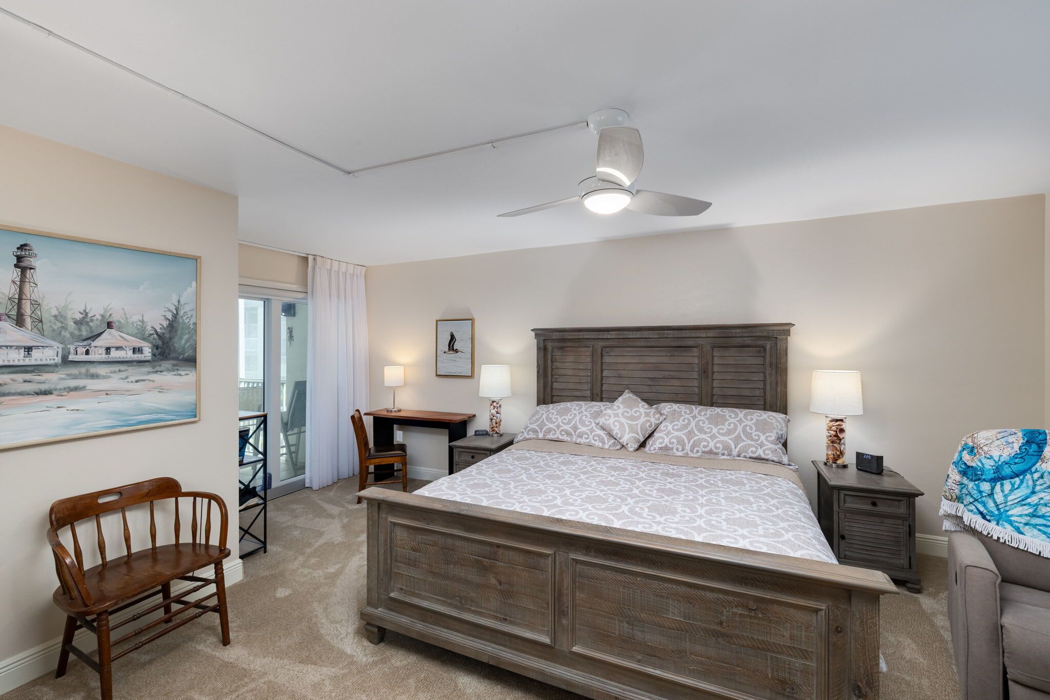 A bedroom with a large bed, a desk, a chair, a ceiling fan, and a painting on the wall.
