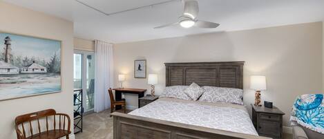 A bedroom with a large bed, a desk, a chair, a ceiling fan, and a painting on the wall.