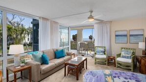 A spacious living room with a large window, a ceiling fan, a sofa with cushions, a coffee table, and two wicker chairs. - Island Beach Club 240F | Exquisite Oceanfront Residence | Sun, Sea and Sand (Sanibel)