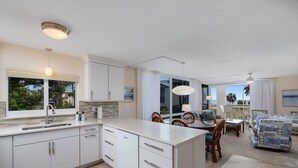A modern kitchen with white cabinets, a large island, and a dining area with wooden chairs and a table. - Island Beach Club 240F | Exquisite Oceanfront Residence | Sun, Sea and Sand (Sanibel)
