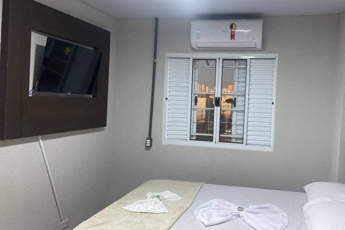 Habitación de hotel con cama, ventana con persianas, equipo de aire acondicionado y televisor montado en la pared.