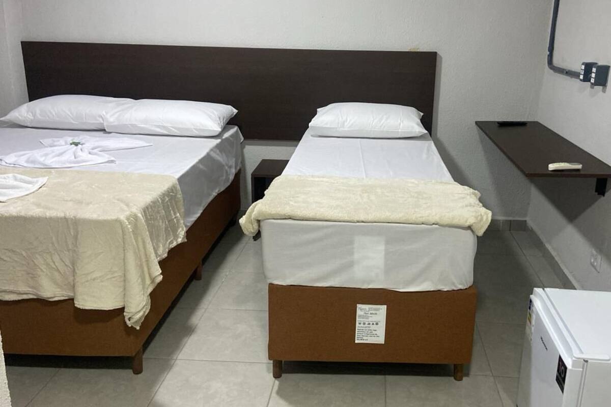 Dos camas individuales con sábanas blancas, una mesita de noche pequeña y un refrigerador blanco.