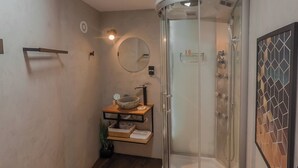 A bathroom with a glass shower enclosure, a white toilet, and a wooden vanity with a round mirror.