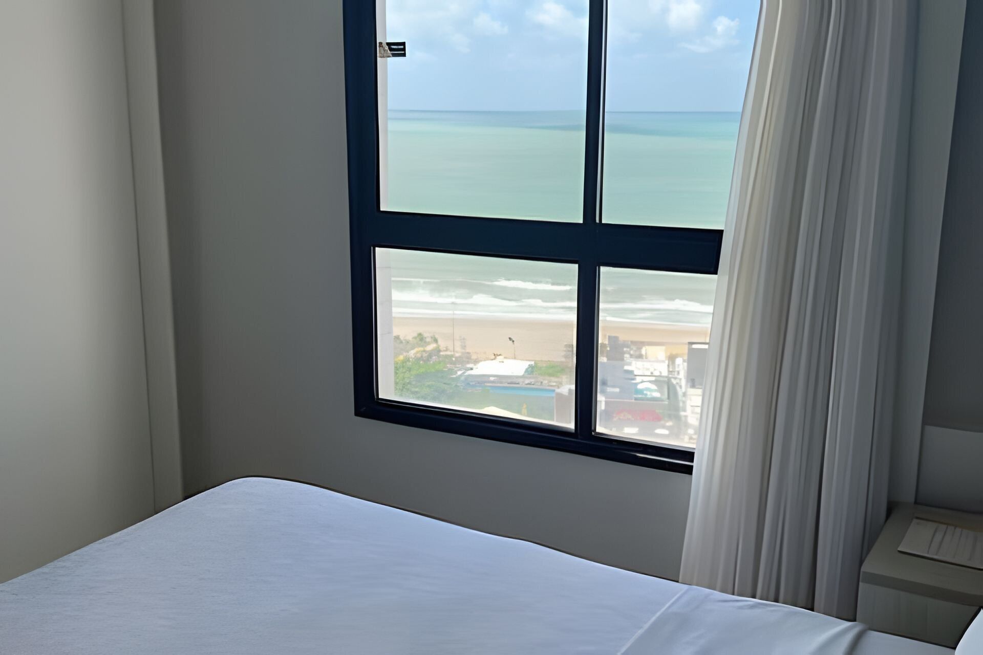 Quarto de hotel com uma janela grande que oferece vista para a praia e o oceano.