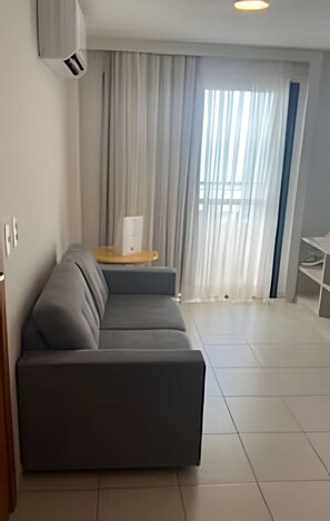 A modern living room with a grey sectional sofa, a wooden side table, and a large window with sheer curtains. - Mar e Descanso  (Natal)