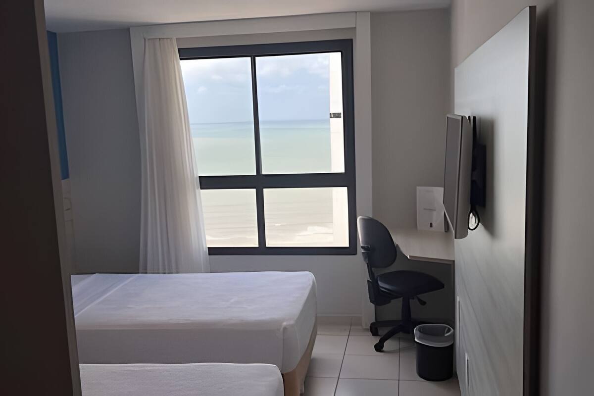 A hotel room with two beds, a desk, a chair, a TV, and a large window with a view of the beach.