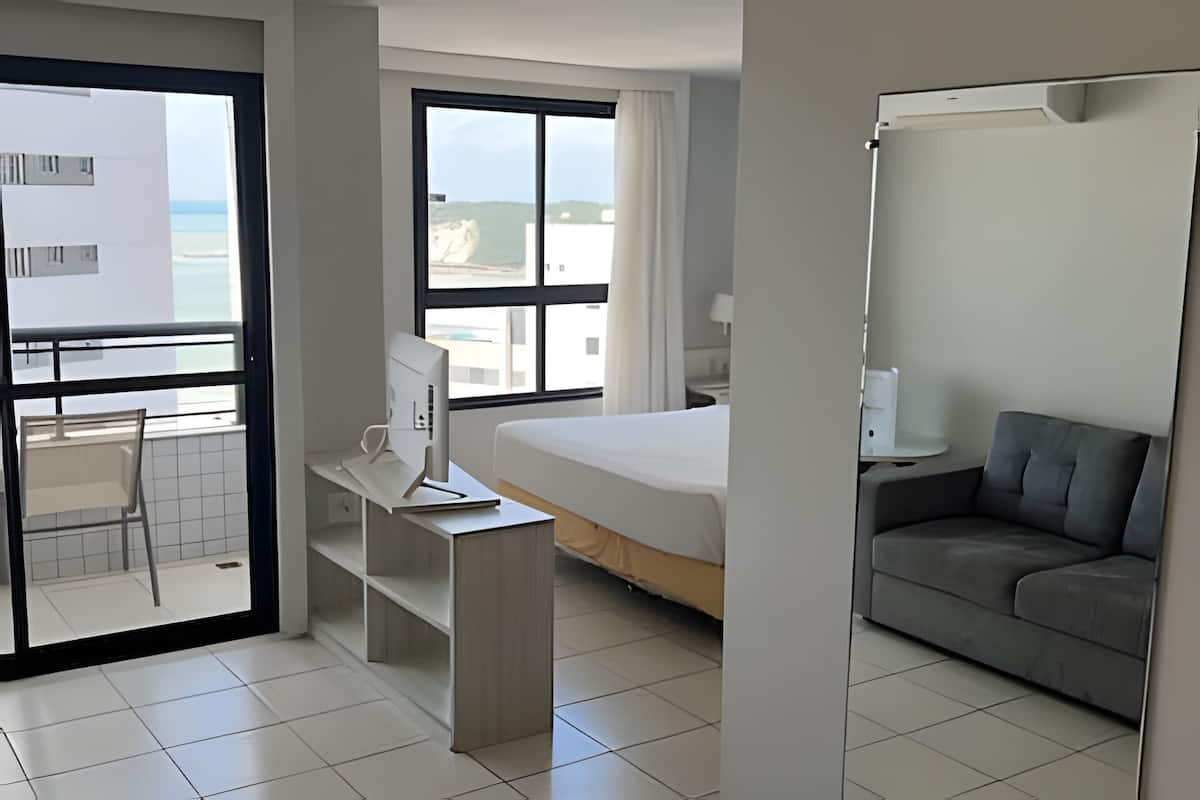 Een hotelkamer met een bed, een bureau met een computer, een sofa, een balkon met uitzicht op zee en een raam met gordijnen.