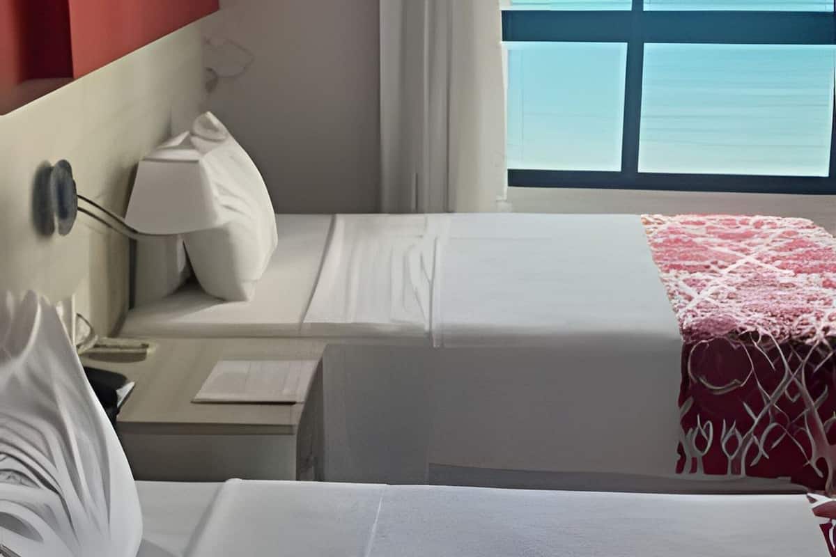 A hotel room with two beds, a nightstand, a lamp, a red and white patterned throw, and a view of the ocean.