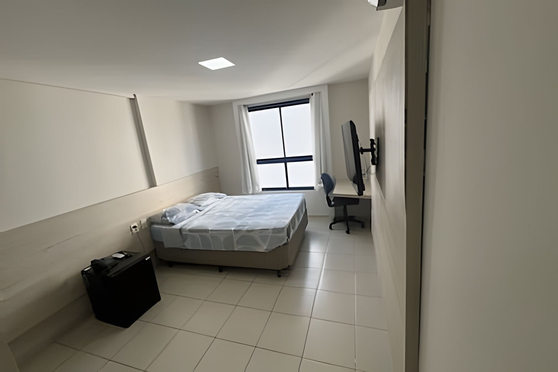 Um quarto com uma cama, uma escrivaninha, uma cadeira, um computador e uma janela.