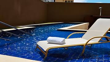 Piscina no terraço com espreguiçadeira e toalha, cercada por um muro marrom e um céu claro.