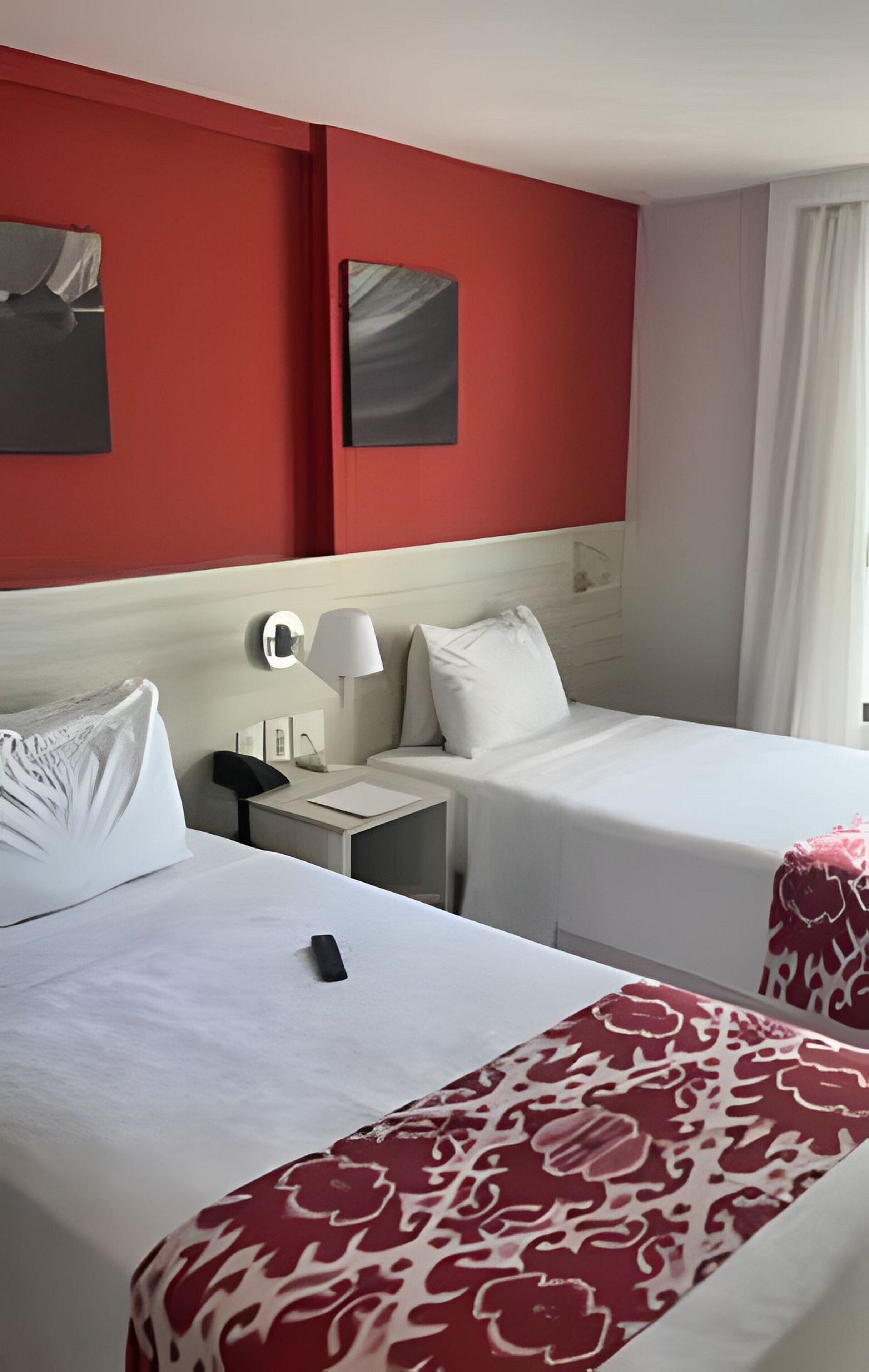 Habitación de hotel con dos camas, una mesita de noche con lámpara, un reloj de pared y una ventana con cortinas.