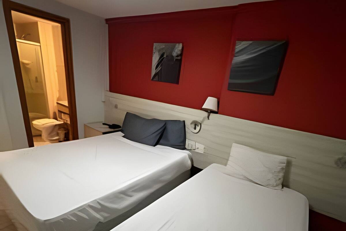 Een hotelkamer met twee bedden, een rode accentmuur, een nachtkastje met een lamp en een zichtbare badkamer door een geopende deur.